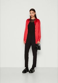 Chemise rouge en satin à boutons, superposée à un top en mesh noir, associée à un pantalon noir et des chaussures noires imposantes. Petit sac à main en accessoire.
