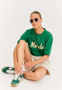 Žena sedí prekrížene v nohách, oblečená v zelenom tričku s nápisom "Merde", pruhovaných kraťasoch, okrúhlych slnečných okuliaroch, farebnom náhrdelníku a zelených teniskách s ružovými akcentmi.