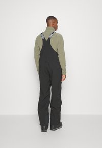 Black overalls with adjustable straps and a back pocket. Worn over a gray sweatshirt. Textured fabric with a smooth finish.