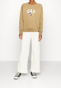 Beżowy sweter z białym napisem "GAP" i grafikami w stylu kreskówkowym. Połączony z szerokimi białymi spodniami i czarnymi trampkami za kostkę.
