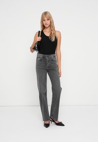 Jean taille haute gris avec jambe droite, comportant des poches avant et une fermeture éclair. Associé à un haut sans manches noir et des talons noirs.