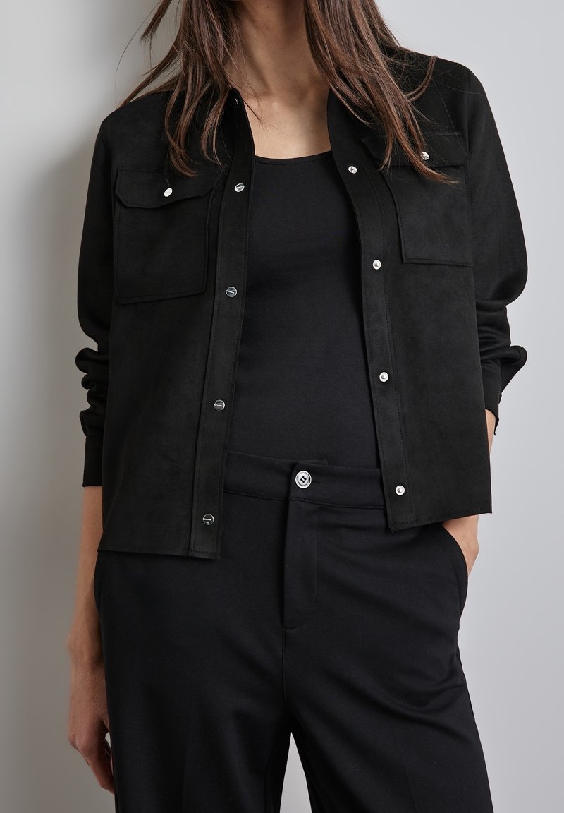 Svart mockaskjorta med två bröstfickor, tryckknappar och en avslappnad passform, kombinerad med en svart topp och byxor.