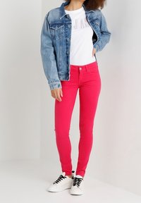 Denimjacke, helle Waschung, Knopfverschluss; weißes T-Shirt mit "JEANS"-Text; leuchtend pinkfarbene Skinny-Jeans; weiße Sneakers mit schwarzen Schnürsenkeln.