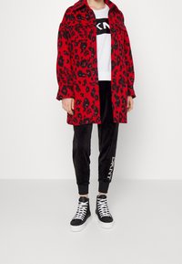 Röd leopardmönstrad oversized kappa, vit grafisk t-shirt, svarta velourbyxor med "DKNY" text, och svarta högklackade sneakers med vita sn ören.