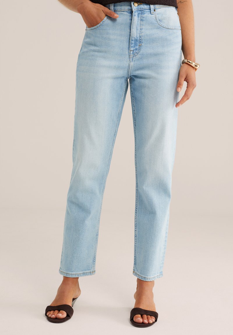 Hellblaue, hoch taillierte Jeans mit geradem Bein, getragen mit schwarzen Sandalen mit offenem Zeh und einem goldenen Armband am rechten Handgelenk.