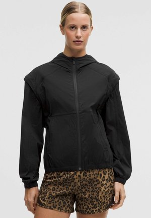 Nuori nainen, jolla on musta vetoketjullinen huppari ja ruskeat leopardikuvioiset shortsit, seisoo yksinkertaisen vaalean taustan edessä.