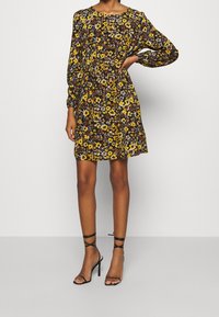 Robe fleurie sur fond noir avec des fleurs jaunes et bleues, manches longues, taille cintrée et coupe décontractée, associée à des talons noirs à lanières.