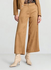 Pantalon large couleur brun clair en matière douce semblable au daim, avec une texture lisse et un léger éclat. Associé à des talons beiges à bout pointu.