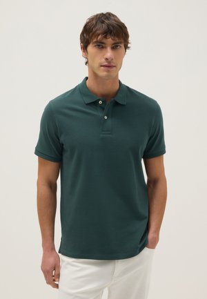 Hombre vestido con camiseta polo de manga corta verde oscuro y pantalones blancos, de pie contra un fondo claro y liso con una mano en el bolsillo.