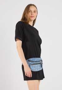 Tricou negru cu mânecă scurtă și dantelă, purtat cu o borsetă texturată albastră, având două buzunare cu fermoar și o formă rotunjită.