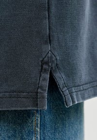 Ourlet en tissu gris foncé texturé avec une fente latérale au-dessus d'un jean en denim bleu, montrant un gros plan du textile et des coutures.