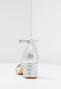 Silver heeled sandal with ankle strap, smooth metallic finish, and a braided-textured block heel. Side view showcases design details.