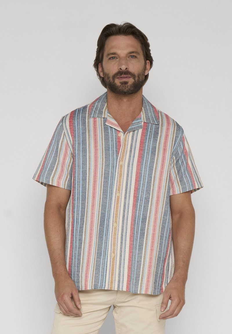 Homme aux cheveux foncés et à la barbe portant une chemise à manches courtes à boutons avec des rayures verticales rouges, bleues, blanches et beiges, debout devant un fond uni.