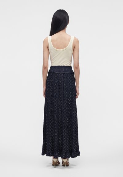 Femme de dos, portant un haut beige sans manches, une longue jupe sombre à motifs et des talons, debout sur un fond uni et clair.