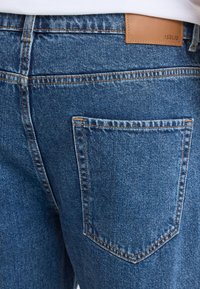 Gros plan d'une poche arrière de jean bleu avec des surpiqûres orange et un patch en cuir marron portant la marque « !SOLID » sur la ceinture.