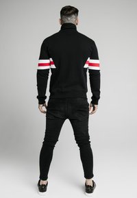 Sudadera negra con cuello alto, que presenta rayas verticales rojas y blancas en las mangas. Combinada con pantalones skinny negros y zapatillas negras.