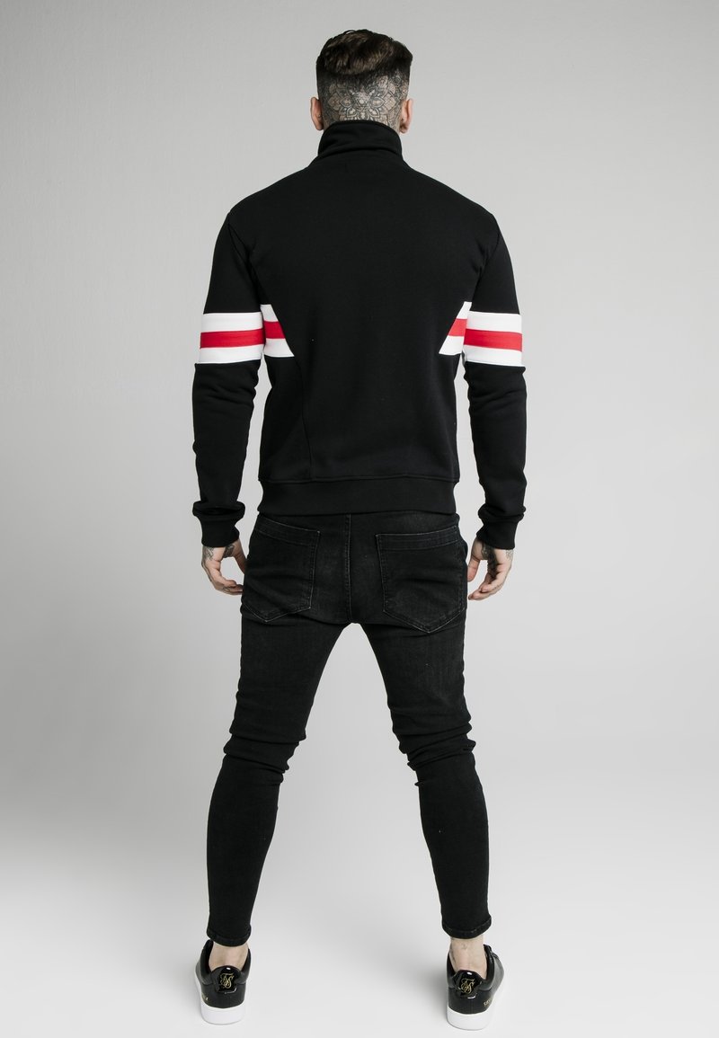 Sudadera negra con cuello alto, que presenta rayas verticales rojas y blancas en las mangas. Combinada con pantalones skinny negros y zapatillas negras.