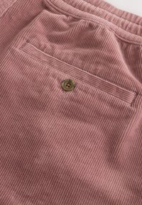 Gros plan sur un tissu en velours côtelé rose poussiéreux avec une poche arrière munie d’un bouton marron et des coutures visibles.