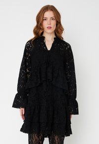 Rochie din dantelă neagră, cu un design stratificat, mâneci lungi în formă de clopot, decolteu în V și accente volanate. Modele florale intricate pe întreaga suprafată.