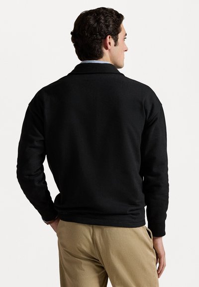 Hanorac negru cu un design simplu, manșete ribbate și guler rotund, purtat împreună cu pantaloni bej. Materialul pare moale și texturat.