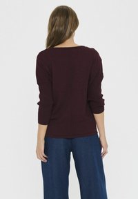 Burgundijska pletena majica ohlapnega kroja s tremi četrtinami rokavov in osrednjim šivom, ujemajoča se z širokimi denim hlačami. Vidna teksturirana tkanina.