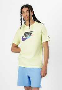 Nike majica u svijetlozelenoj boji, s velikim višebojnim dizajnom logotipa. Uparena s plavim kratkim hlačama, kratkim rukavima i ležernim krojem.