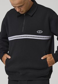Sudadera negra con cremallera y cuello, con una franja horizontal blanca y estampada en el pecho y bolsillos laterales.