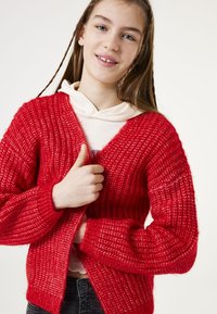 Rode gebreide cardigan met een open voorkant, een gestructureerd oppervlak, geribbelde mouwen en een losse pasvorm. Gedragen over een lichtgekleurde hoodie.