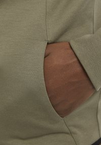 Olive green sweatshirt fabric with a side pocket. Close-up of a hand against textured material, highlighting seam and pocket detail.