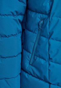 Gros plan sur le tissu d'une veste matelassée bleue avec une poche zippée verticale sur le côté droit.
