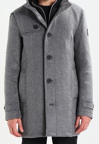 Šedý kabát v herringbone vzoru s vysokým límcem, zapínáním na knoflíky a bočními kapsami. Má jemnou texturu a přiléhavý střih.