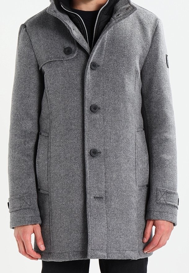 Šedý kabát v herringbone vzoru s vysokým límcem, zapínáním na knoflíky a bočními kapsami. Má jemnou texturu a přiléhavý střih.
