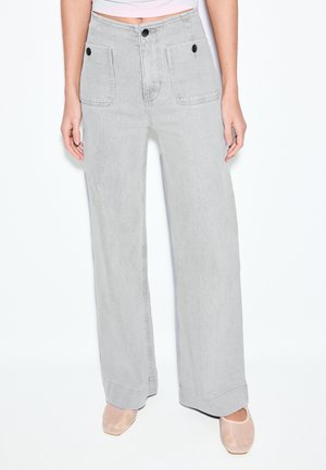 Hellgraue, hoch taillierte, weit geschnittene Hosen mit großen Vordertaschen und schwarzen Knöpfen, getragen mit blassrosa Ballerinas und einem kurz geschnittenen Oberteil.