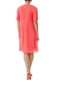 Robe plissée en corail vif, à manches courtes, décolleté arrondi, design fluide, longueur au-dessus du genou, accompagnée de talons roses assortis.