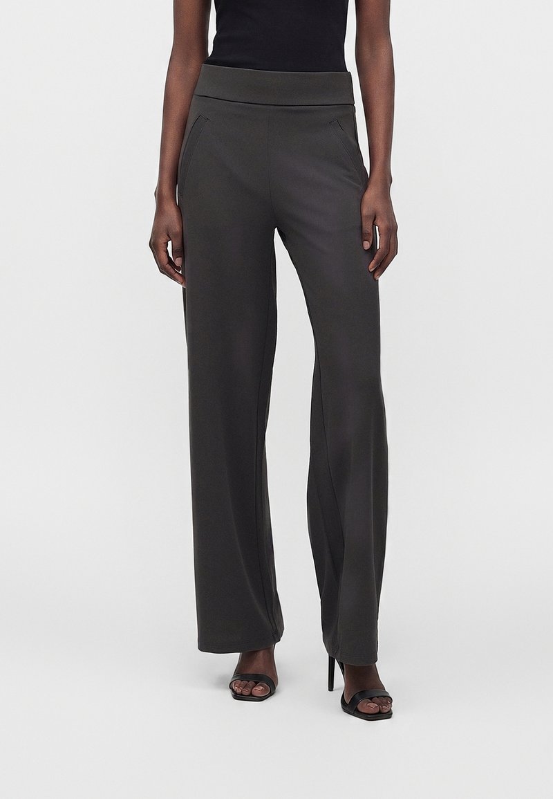 Femme portant un pantalon noir taille haute à jambes larges et des sandales noires à talons ouvertes, debout sur un fond uni.