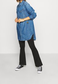 Camicia di jeans blu medio con maniche lunghe, chiusura a bottoni davanti, due tasche sul petto e orlo curvo, abbinata a pantaloni flare neri e sneaker.