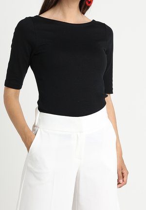 Kvinde iført en tætsiddende sort top med ærmer til albuen og højtaljede, løstsiddende hvide bukser, stående med den ene hånd i lommen.