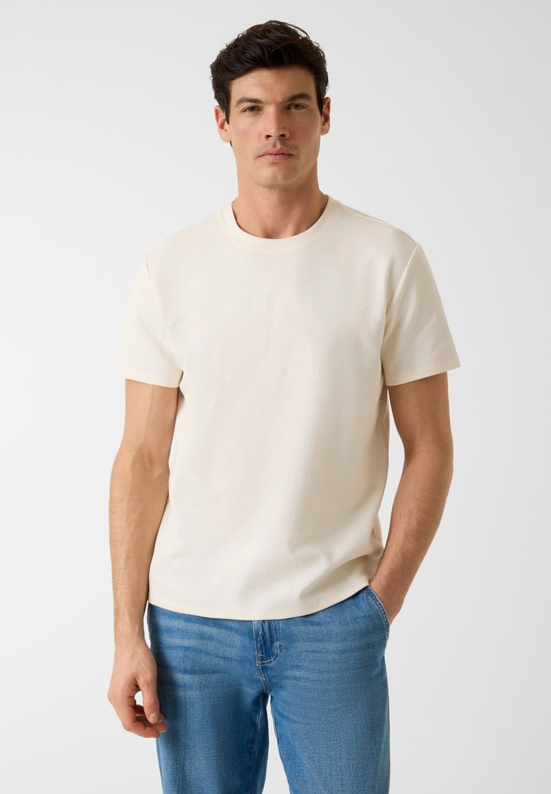 Homme aux cheveux foncés portant un t-shirt crème à manches courtes et un jean bleu, debout devant un fond clair uni avec une main dans la poche.