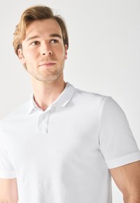 Fehér polo ing struktúrált anyagból, két gombos gallérral és rövid ujjakkal. Egyszerű dizájn, minta vagy díszítés nélkül.