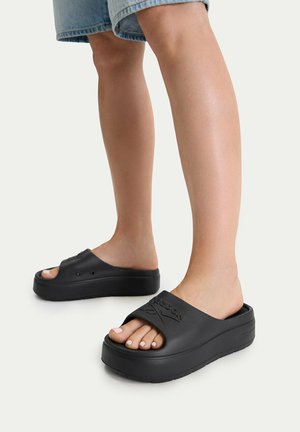 Beine mit schwarzen Plateau-Slide-Sandalen mit offenen Zehen und einem Reebok-Logo, kombiniert mit blauen Jeansshorts vor einem einfarbigen Hintergrund.