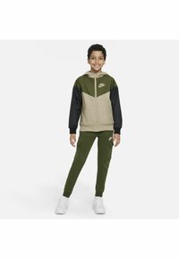 Chaqueta de Nike en beige, verde oliva y negro, con capucha y cierre de cremallera. Combinada con joggers verde oliva que tienen bolsillos tipo cargo. Zapatillas blancas.