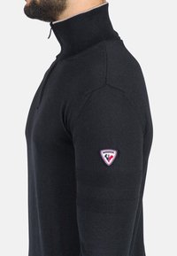 Pullover nero a maniche lunghe con colletto alto, texture a coste e patch del logo Rossignol sulla manica sinistra. Dettaglio con zip al collo.