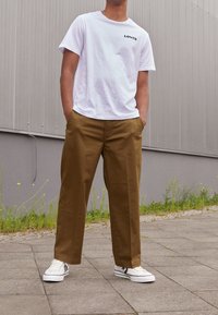 Bílý bavlněný tričko s černým logem "Levi's", spojené s hnědými bavlněnými kalhotami a bílými teniskami s černými akcenty.