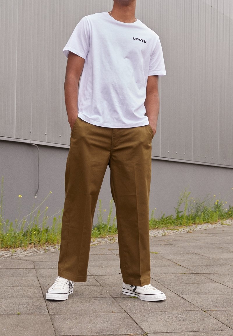 Bílý bavlněný tričko s černým logem "Levi's", spojené s hnědými bavlněnými kalhotami a bílými teniskami s černými akcenty.
