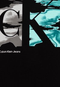 Camiseta negra con un gran logo "CK" en negrita en tonos gris y turquesa, con el texto "Calvin Klein Jeans" en blanco debajo. Textura suave.
