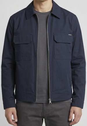 Man draagt een marineblauwe jas met rits en twee klepzakken op de borst, over een donkergrijs T-shirt en een broek.