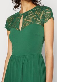 Zielona sukienka z koronkowymi krótkimi rękawami i detalem w kształcie łezki przy dekolcie, z dopasowaną górą i rozkloszowanym dołem. Gładka tekstura.