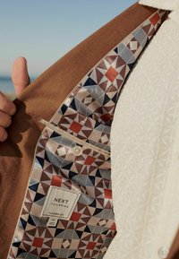 Braun maßgeschneiderte Jacke mit einem geometrischen Futter in Rot, Blau und Creme. Verfügt über einen Knopf und ein Etikett mit der Aufschrift "NEXT Tailoring."