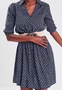 Femme portant une robe bleu marine à petit motif floral, une large ceinture beige et des manches retroussées, posant devant un fond uni.