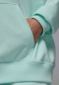 Sudadera verde menta con un bolsillo tipo canguro, puños acanalados y tejido texturizado. El diseño es simple, con costuras suaves y detalles mínimos.